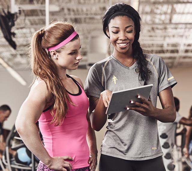 A female personal trainer at Gold's Gym, smiling as she shares fitness insights with a gym member using a tablet.