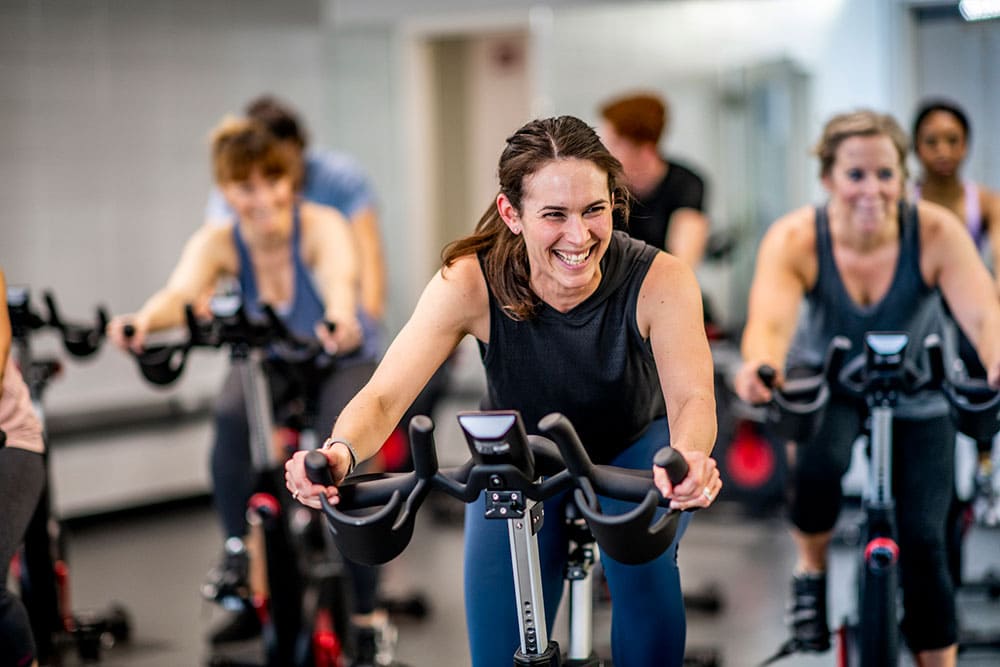 A group of focused women participating in an indoor cycling class, with the foreground featuring a cheerful woman leading the exercise on a stationary bike